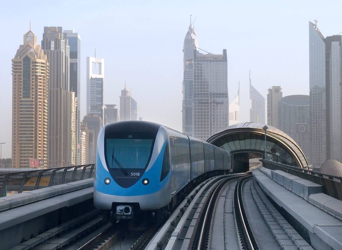 A Dubai Metro train on an elevated guideway with downtown towers behind it.