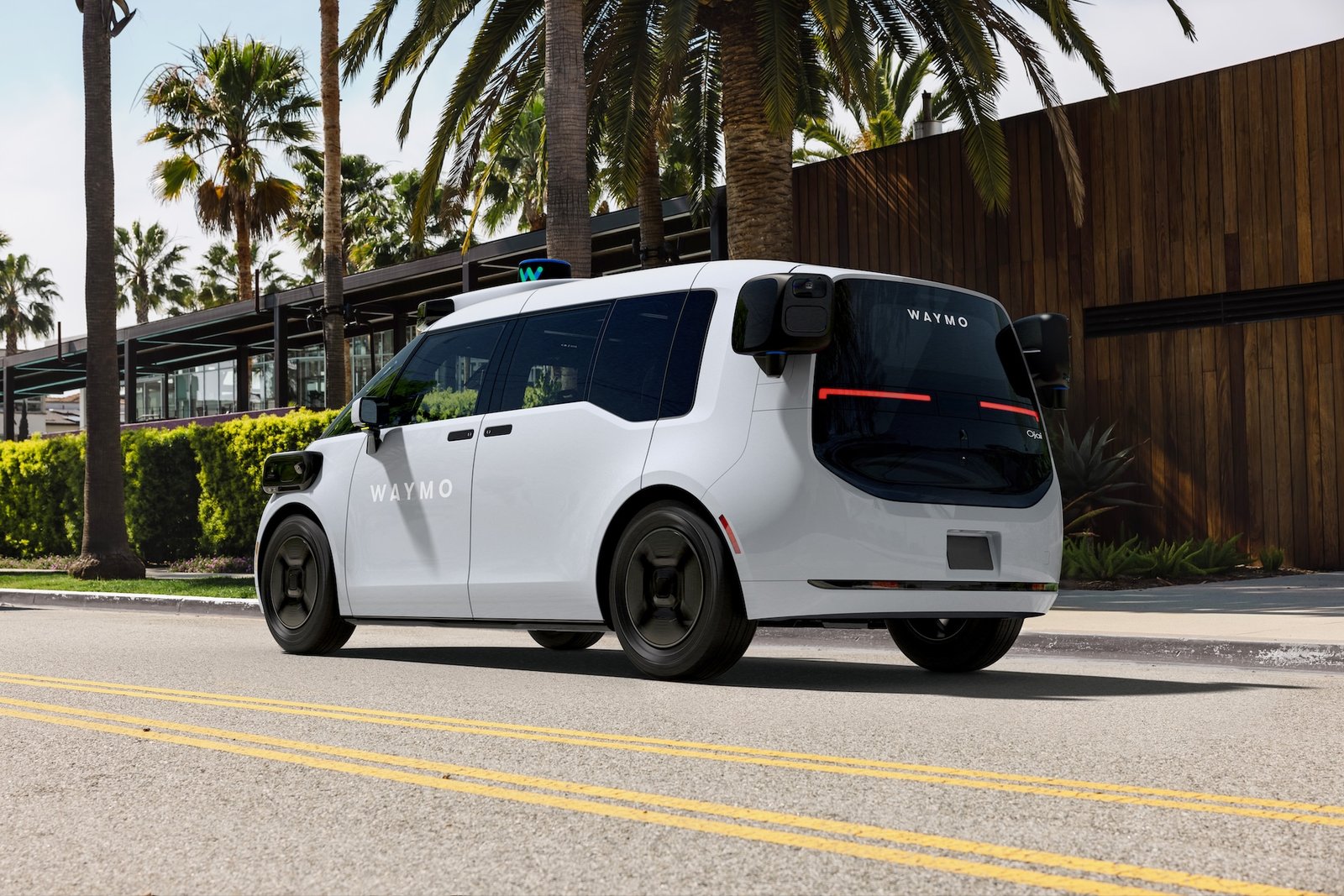 A white Waymo Ojai robotaxi on a palm-lined street, with no driver in the front seat.