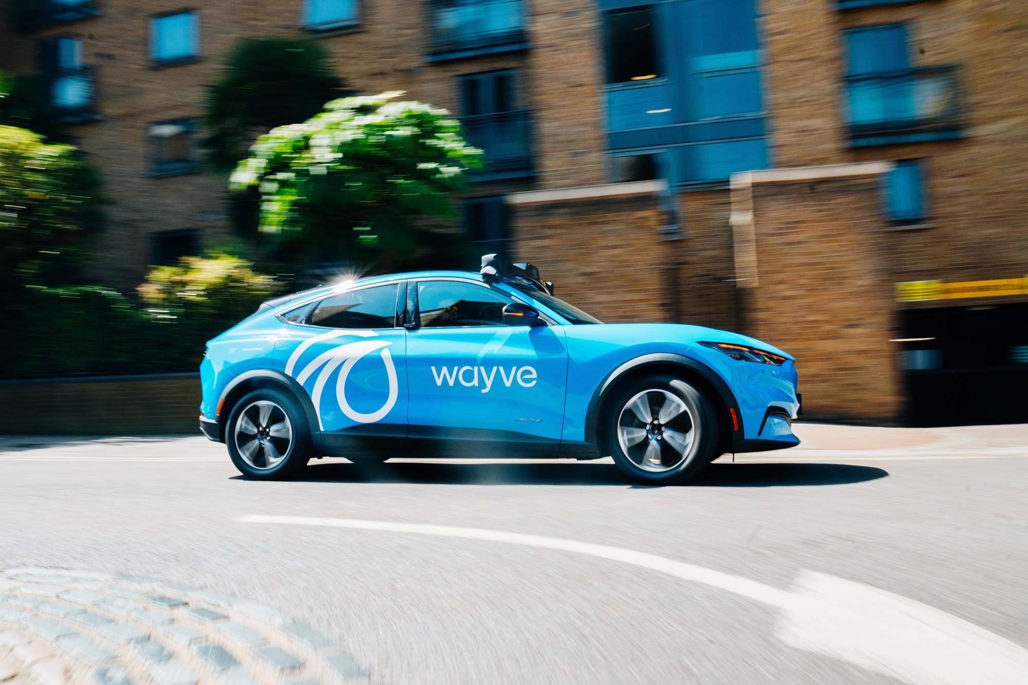 A blue Wayve-branded autonomous vehicle on a city street.